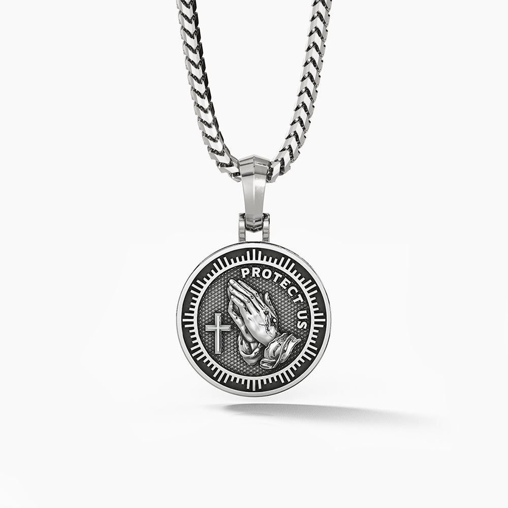 The back of a round Saint Christopher medallion showing sculpted praying hands, a cross and the words protect us