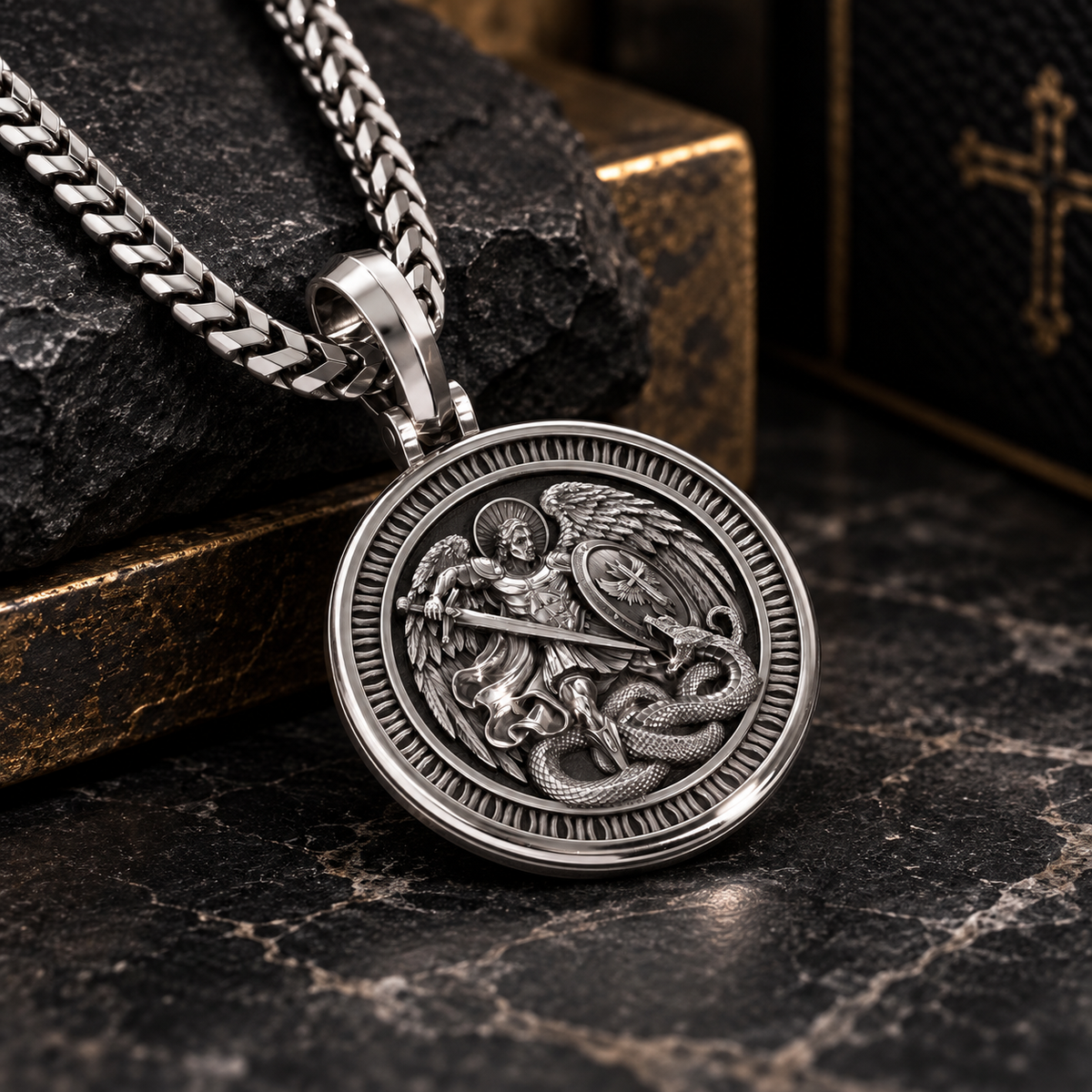 A silver-toned round pendant necklace featuring an embossed Saint Michael the Archangel defeating a dragon design, resting on a dark textured stone surface next to a gold-trimmed wooden box.