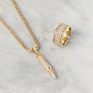a gold and diamond bullet pendant lays next to a mens diamond wedding band on white marble
