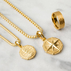 two gold compass pendants lie on white marble, one showing a nautical ship on the back