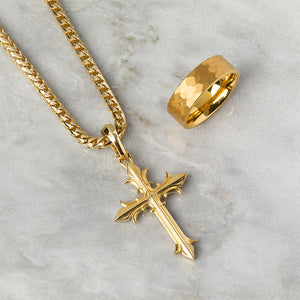 A unique gold necklaces crosses shines as it lies on a white marble surface next to a hammered tungsten ring
