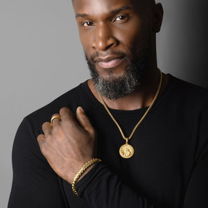 a muscular man in a black shirt rests his hand on his shoulder while wearing a gold Saint Christopher necklace