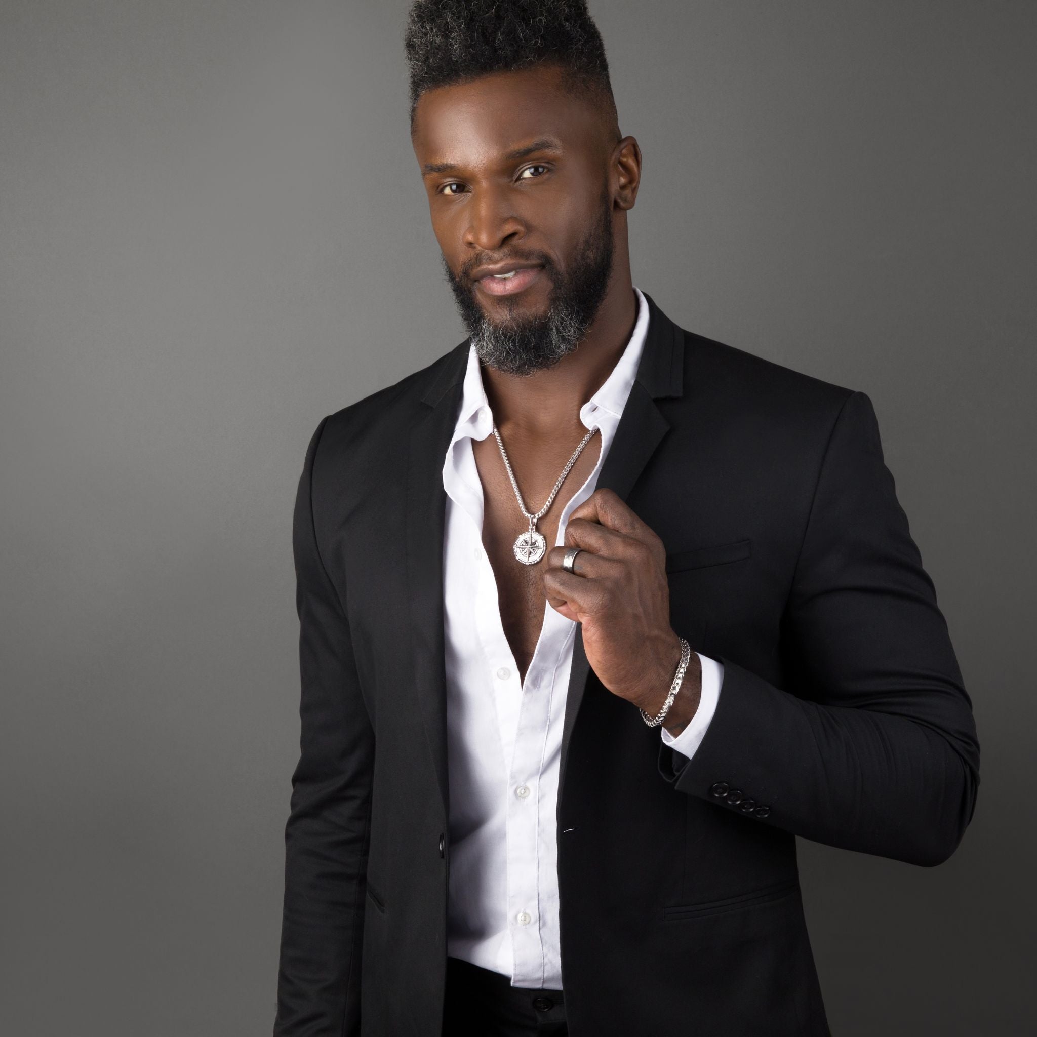 a tall man wearing a black suit grabs his lapel and a silver franco bracelet, tungsten ring, and mens chain shine