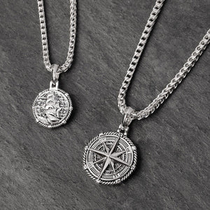 two silver compass pendants lie on slate, one showing a nautical ship on the back