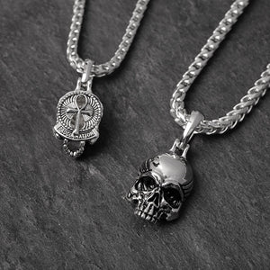 two silver skull pendants lying on slate. One has an ankh on the back